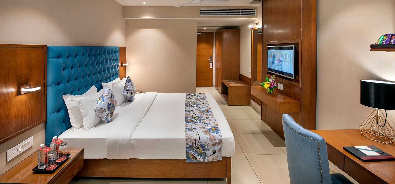 A comfortable hotel room  with a double bed with a vibrant blue headboard, a work desk with a blue chair, and a mounted television - SK Lords Inn Ahmedabad