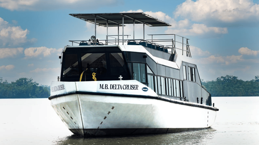 M.B. Delta Cruiser, a white multi-deck cruise boat with shaded upper viewing deck, moored on calm water against a backdrop of green trees, and blue sky.