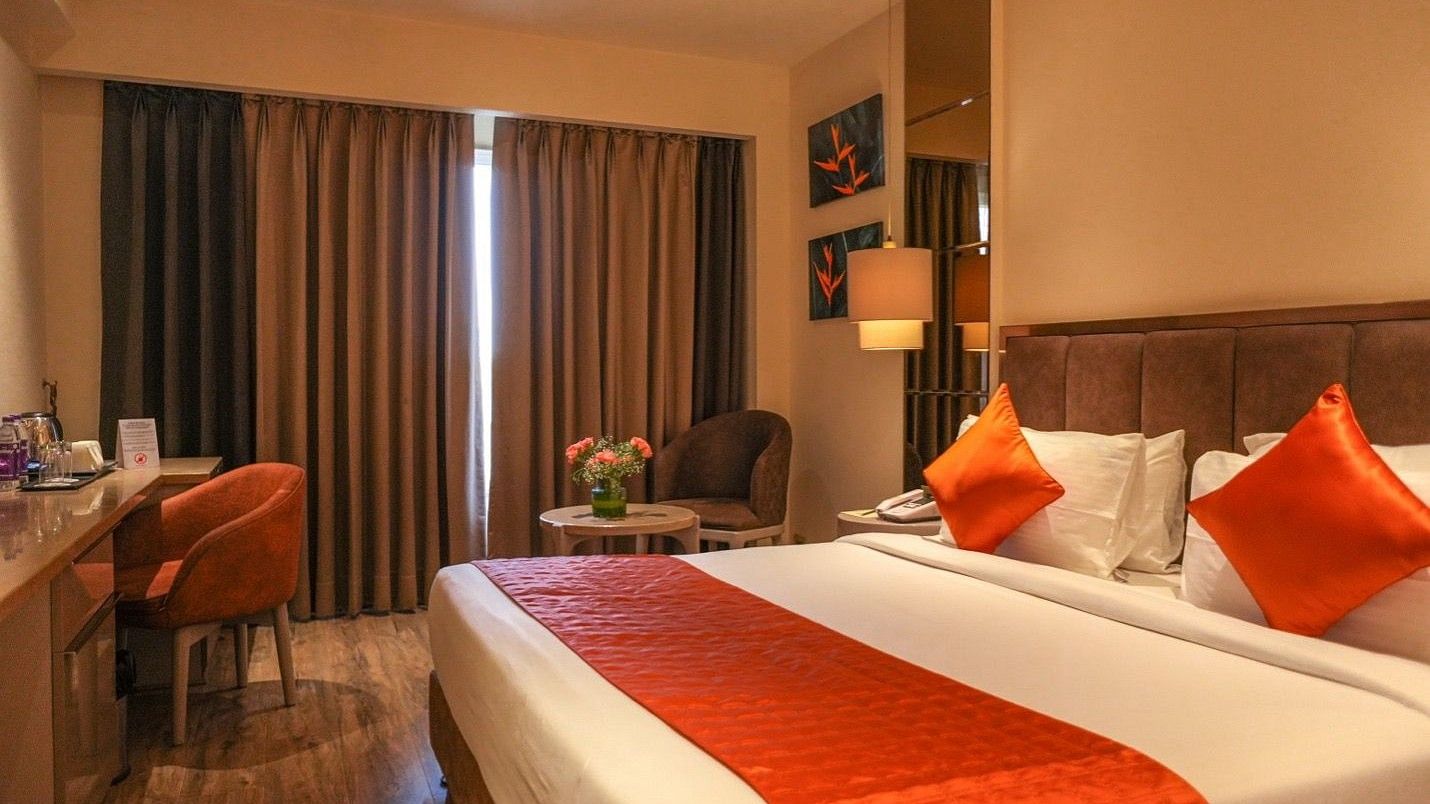 A well-lit, modern guest room at Hotel Saket 27, featuring a cozy double bed with red accents and elegant decor.