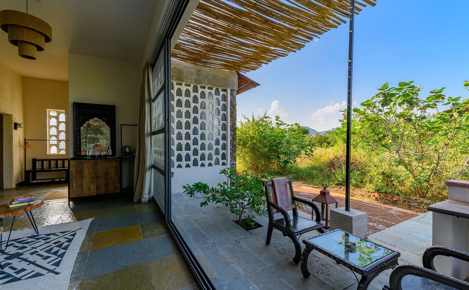 An image of a room with a chandelier, and other decorative items and an expansive glass door over viewing the green landscape - Utsav Camp Sariska