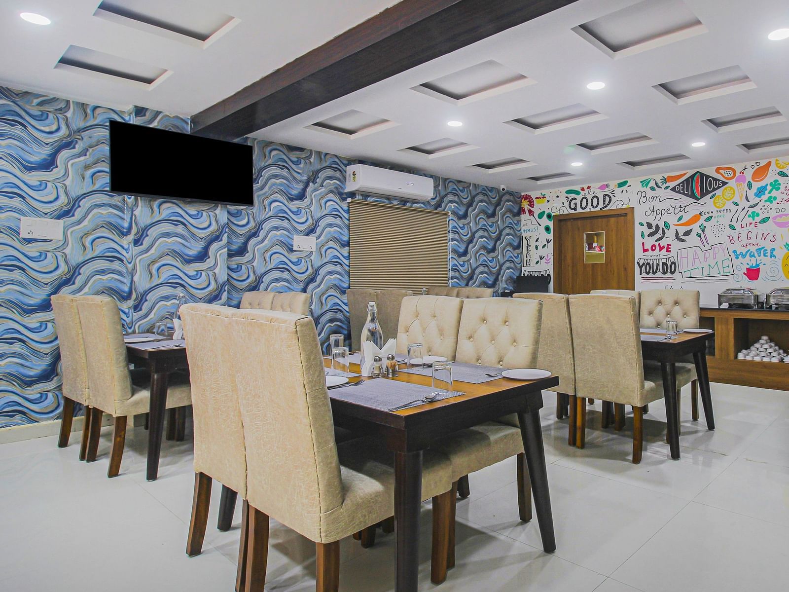 A brightly lit dining space with plush beige chairs, wooden tables and a lively mural wall with inspirational quotes, complemented by a swirling blue wallpaper.