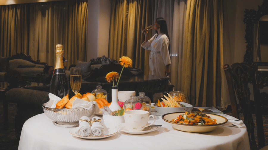 A round dining table set with plated dishes wine glasses and a flower arrangement near drawn curtains at The Suryaa, New Delhi.