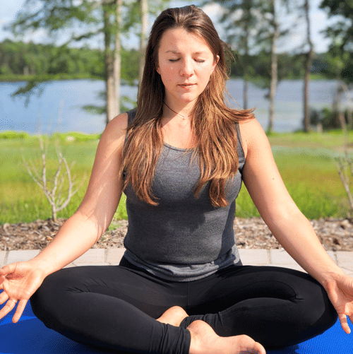 Woman meditating outdoors in a cross-legged pose on a blue yoga mat, with trees and a lake in the background at YO1 Longevity and Health Resorts, Catskills.