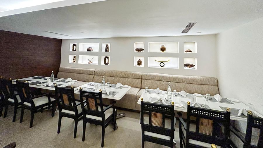 A bright restaurant interior shows set tables with white cloths and dark chairs against a long beige banquette and lit display shelves at The Residency Karur.
