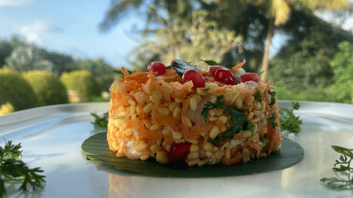 A close-up shot of a sprouted lentil salad, also known as a chaat, garnished with pomegranate seeds and curry leaves, served on a banana leaf on a white plate with a blurred outdoor background of palm trees and greenery.