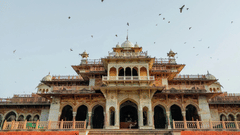 A wide-angle shot of the facade of Albert Hall Museum in Jaipur, featuring its stunning Indo-Saracenic architecture, domes, and pigeons in flight against a bright sky.