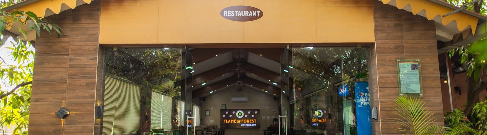 Cosy, glass-fronted restaurant featuring wooden interiors nestled in lush greenery at Daksh Resort and Amusement Park.