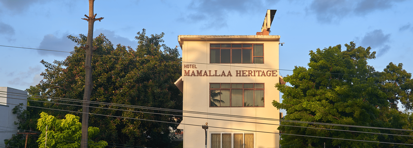 The multi-story exterior of Hotel Mamallaa Heritage, a white building with brown roof accents, set against a clear blue sky and surrounded by some greenery.