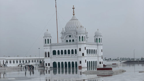 A white marble temple with domes and flags, surrounded by a spacious courtyard covered in white tiles, under a cloudy sky.