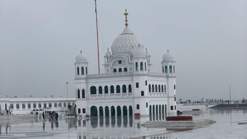 A white marble temple with domes and flags, surrounded by a spacious courtyard covered in white tiles, under a cloudy sky.
