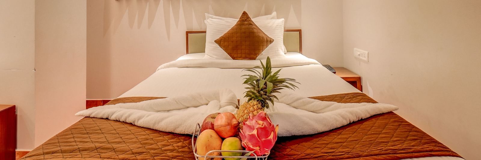 A spacious bed at Harigovinda's JNStay, with neatly arranged blankets, pillows, and a fruit basket, with windows covered by curtains above