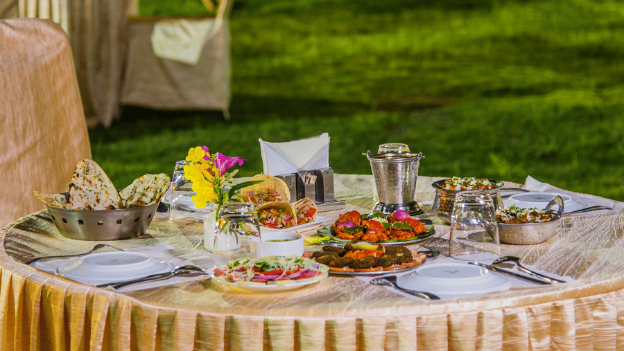 A round dining table covered with a cloth, set with food platters and utensils on a lawn at Garh Govind.