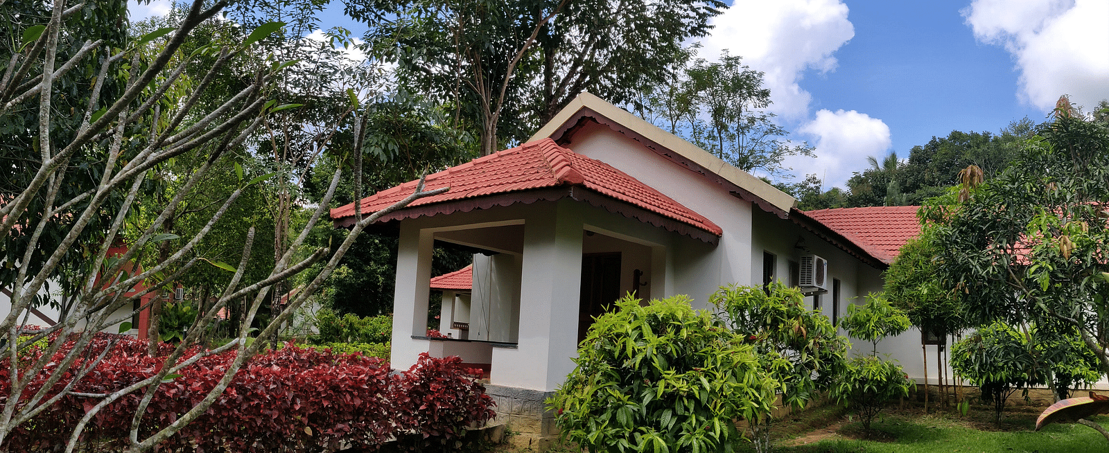 A single-story cottage with a red tiled roof and white walls surrounded by a lush garden with red-flowered shrubs, green lawns, and tall trees against a blue sky with fluffy clouds.