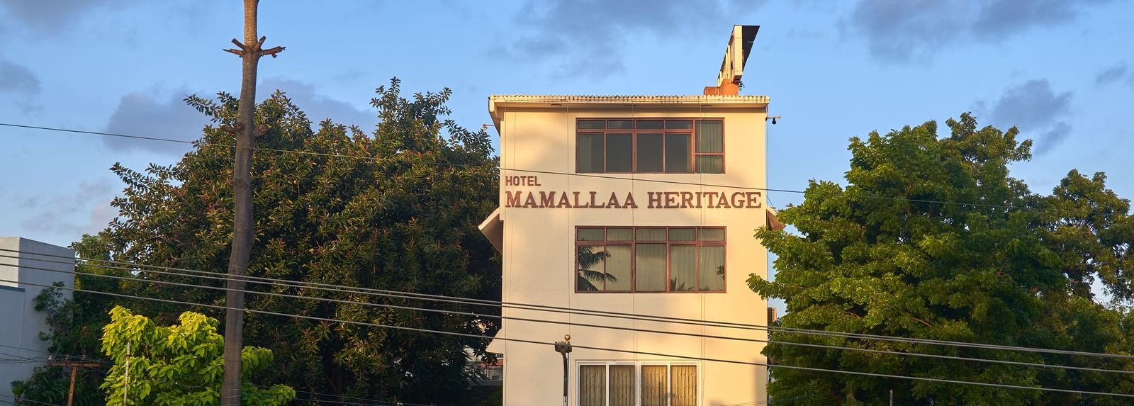 The multi-story exterior of Hotel Mamallaa Heritage, a white building with brown roof accents, set against a clear blue sky and surrounded by some greenery.