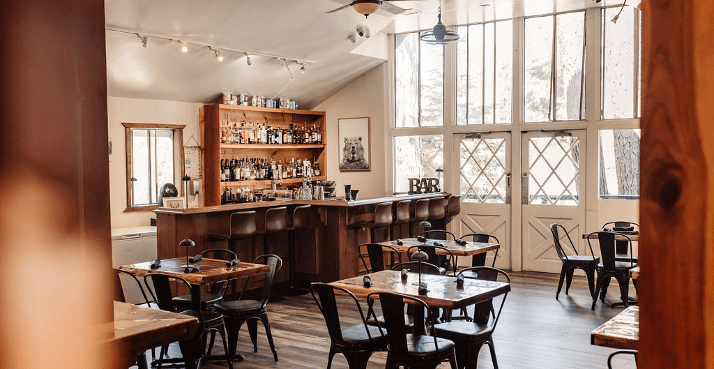 A cafe at Shaver Lake Village Hotel with wooden tables, metal chairs, large windows, and warm lighting, creating an inviting ambiance.