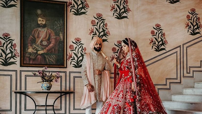 A bride and groom stand together indoors, with a portrait on the wall and floral designs behind them at  Umaid Palace - Best Luxury Heritage Resort Near Jaipur.  