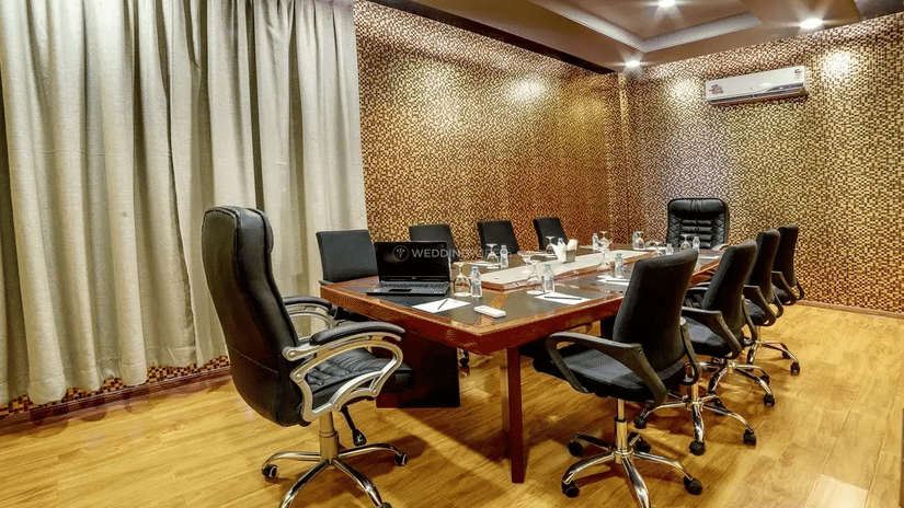 A modern boardroom featuring a long wooden table with black leather chairs, gold patterned wallpaper, and a large neutral-toned curtain at Golden Tulip Kukas, Jaipur.