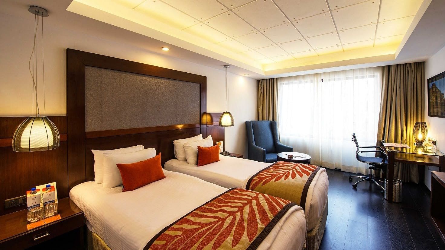 Deluxe Room at The Cunningham Hotel featuring twin beds with decorative runners, warm lighting, pendant lamps, a seating area, and a functional workspace near a large window with natural light