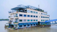 Exterior of Polo Floatel Kolkata, a multi-storey floating hotel moored on the river with large windows, rooftop seating and a blue gangway under an overcast sky.