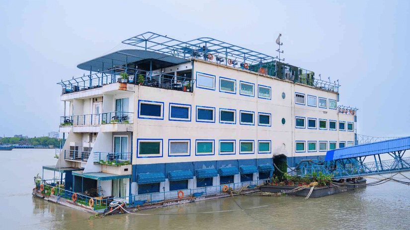 Exterior of Polo Floatel Kolkata, a multi-storey floating hotel moored on the river with large windows, rooftop seating and a blue gangway under an overcast sky.