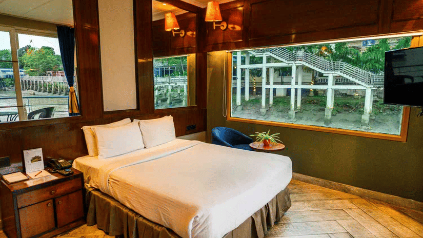 River-view room at Polo Floatel Kolkata with a double bed, warm wood-panelled walls, soft lighting and a large window overlooking the riverside steps and water.