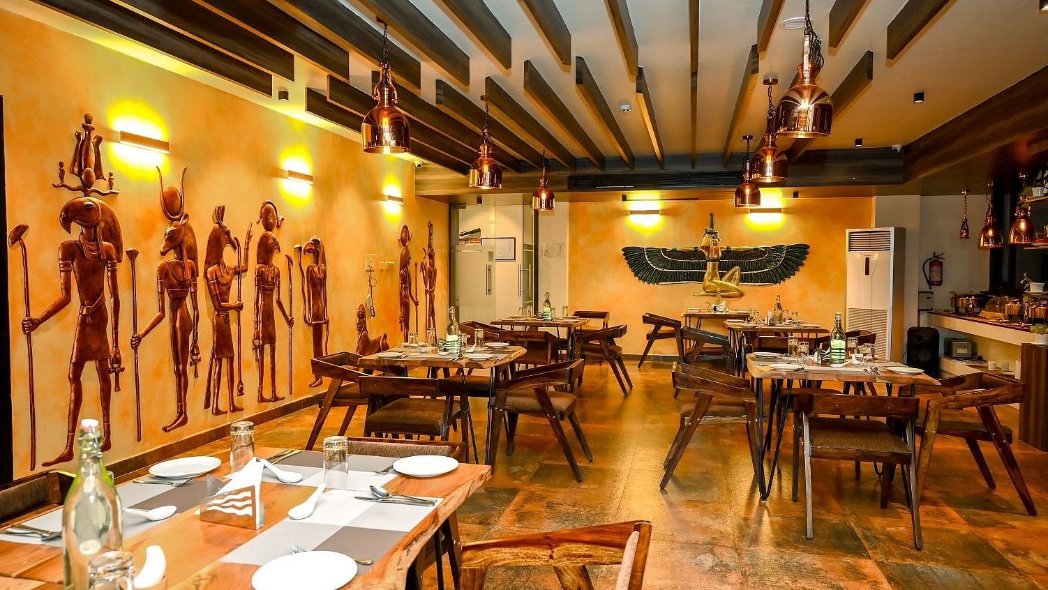 A detailed view of a dining table set for guests in the restaurant, with a focus on the wooden furniture and the warm, ambient lighting from the ceiling fixtures - Sea Hills Hotels, Sri Vijaya Puram (Port Blair)