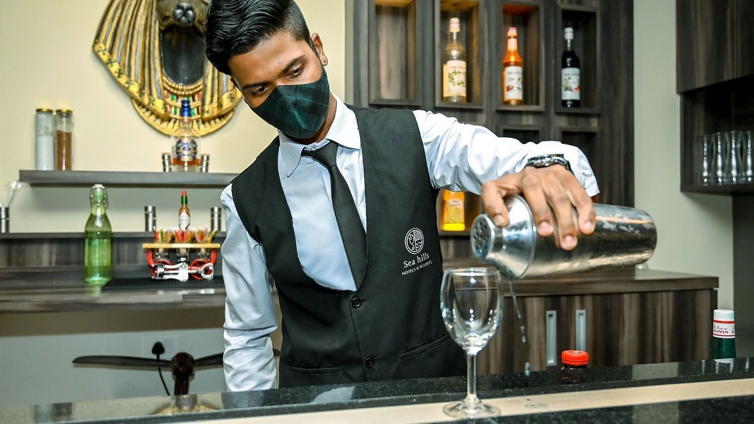 A bartender wearing a face mask and a white shirt with a black tie pours a drink from a bottle into a glass at a well-stocked bar - Sea Hills Hotels, Sri Vijaya Puram (Port Blair)