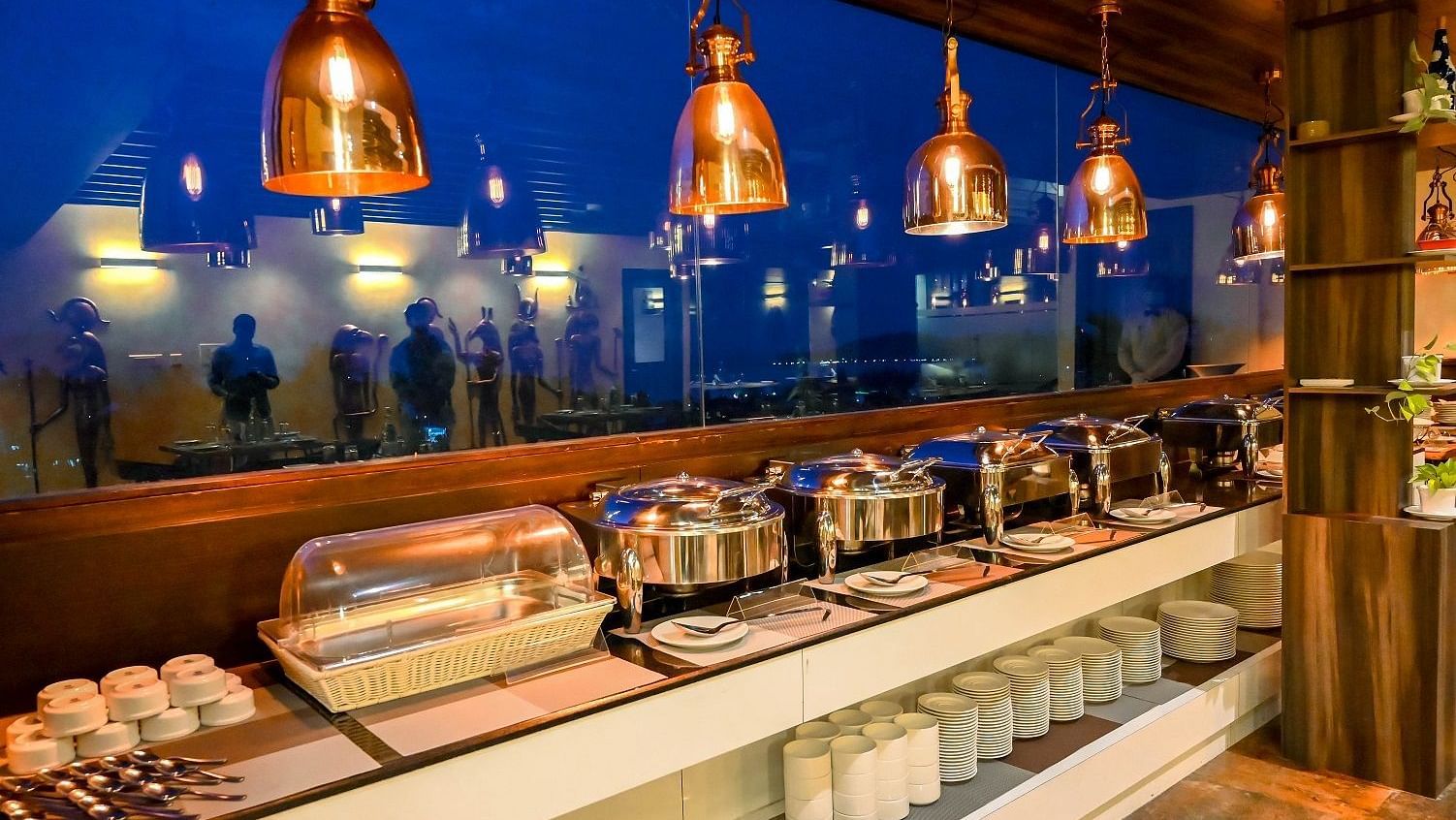 A buffet area in a restaurant, with various dishes in chafing dishes, glasses, and utensils, illuminated by warm hanging lights - Sea Hills Hotels, Sri Vijaya Puram (Port Blair)