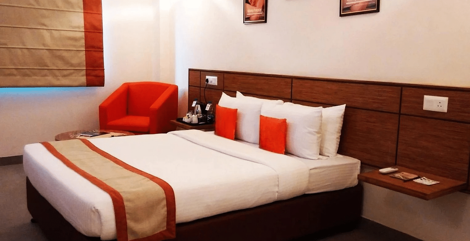 a close up shot of the queen size bed with a small red cushioned arm chair in the room - Max Hotel Jabalpur