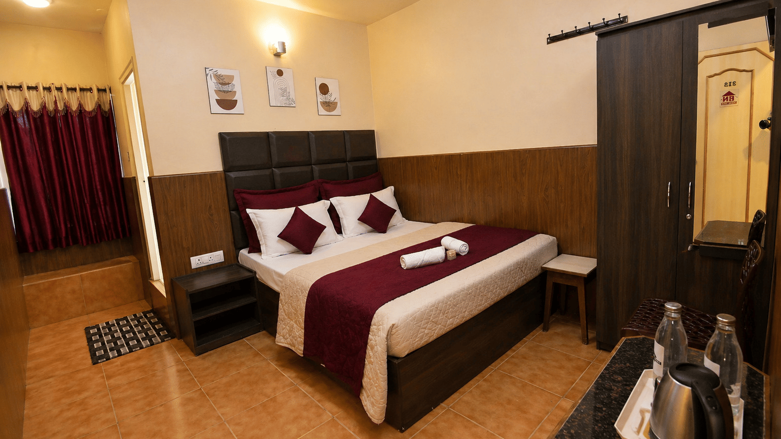 A neatly-made bed with towels, a tall wooden wardrobe, and a small bedside table in the Deluxe Double Room With Extra Cot at BN Tourist Home, Ooty.