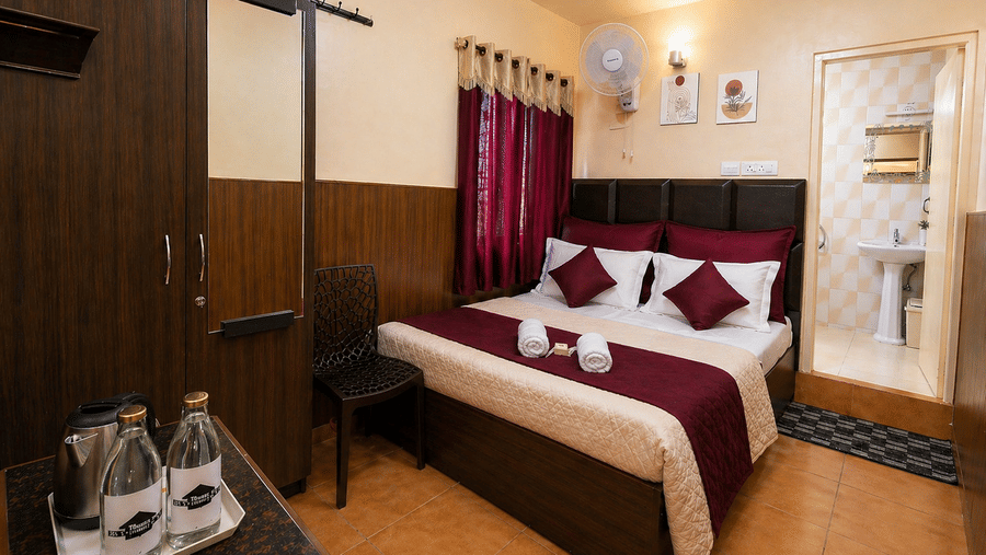 A plush bed next to a large wardrobe with a mirror, a wall-mounted fan, and a kettle setup on a table in the Deluxe Double Room at BN Tourist Home, Ooty.