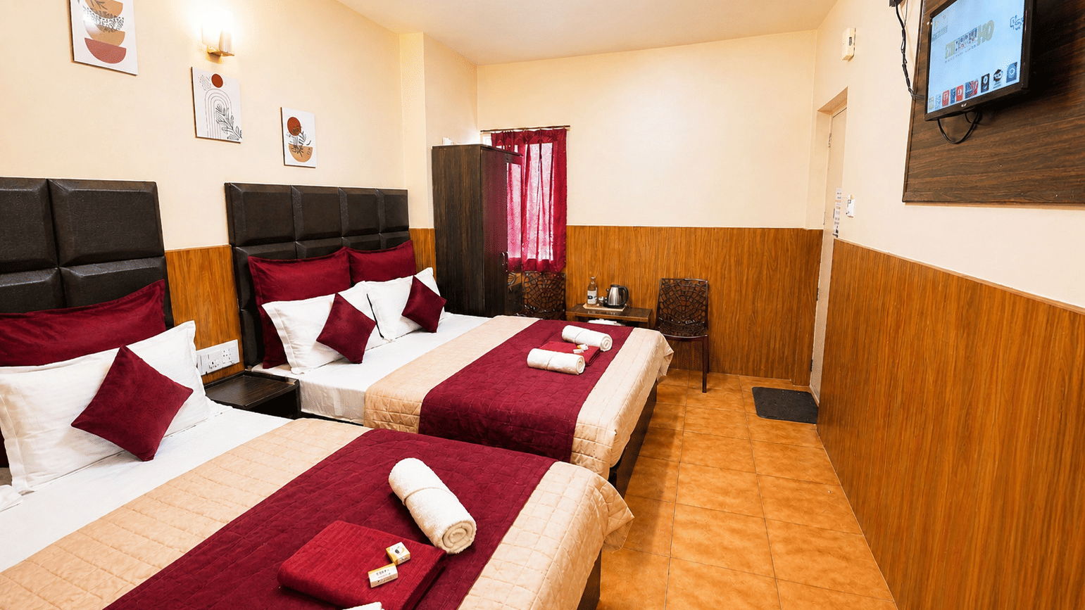 Deluxe Quadruple Room at BN Tourist Home, Ooty, featuring 2 plush beds facing a wall-mounted television screen next to a wardrobe.