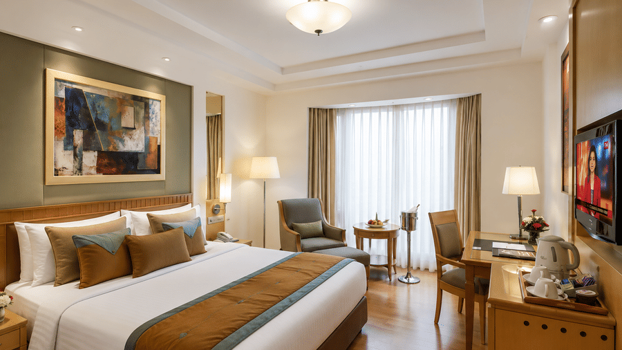 Hotel room with a bed dressed in white and gold linen, a wall-mounted artwork, a seating area with an armchair and a wooden work desk nearby.
