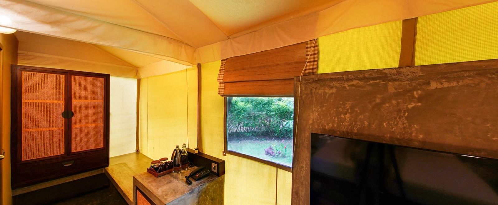 Wooden cabinet, stone counter with bottled water and tray, telephone, and window with garden view inside a yellow fabric-walled tent at Fireflies Resorts, Kabini.