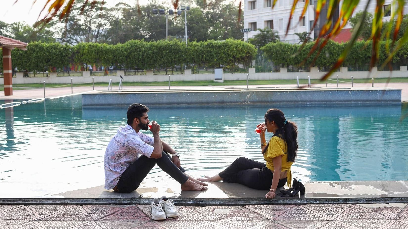 A man and a woman sit on the edge of a pool facing each other, talking and smiling. Trees and buildings are visible in the background at Jenneys Residency