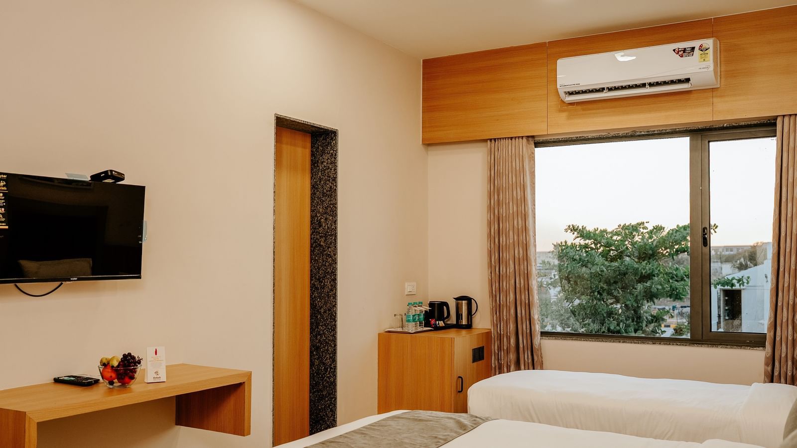 Well-appointed bedroom at Daksh The Grand Gowardhan, Dwarka, showcasing the beds, a wall mounted television, and a window with views.