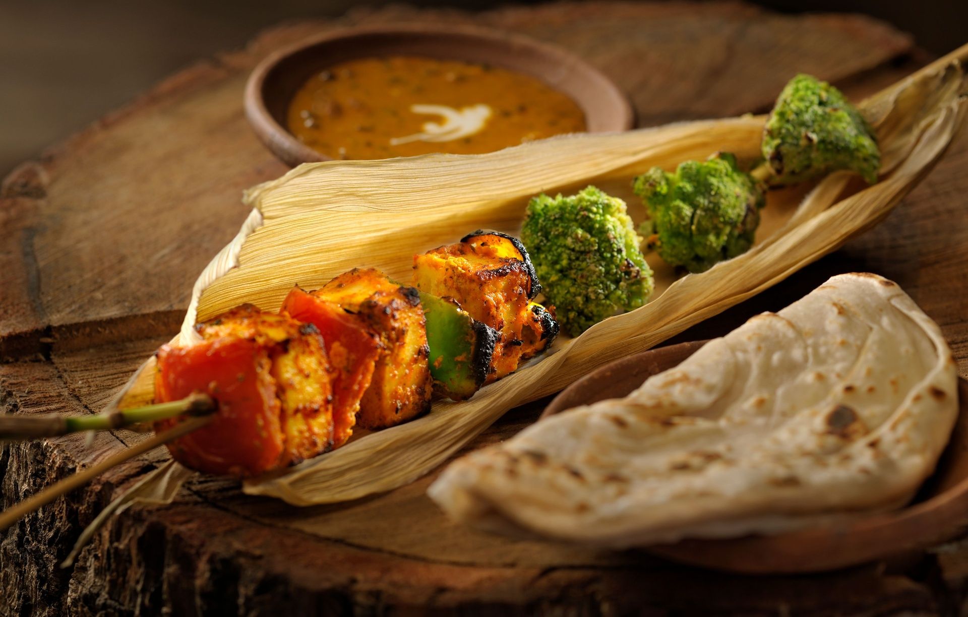 Paneer tikka skewer, broccoli, naan and dal on wood.
