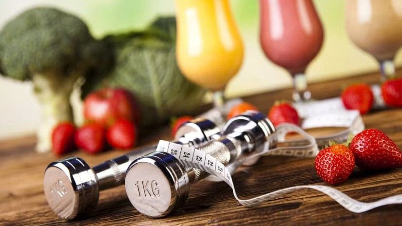 Dumbbells and measuring tape on a table with a variety of fresh smoothies and vegetables in the background, promoting a healthy lifestyle.