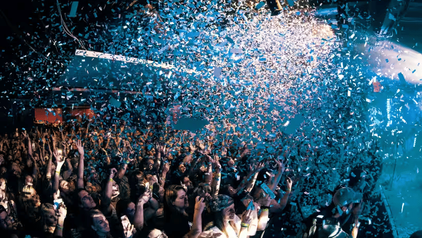 A large crowd of people with hands raised at an indoor party event with confetti falling from above and colorful stage lights illuminating the venue in the background.