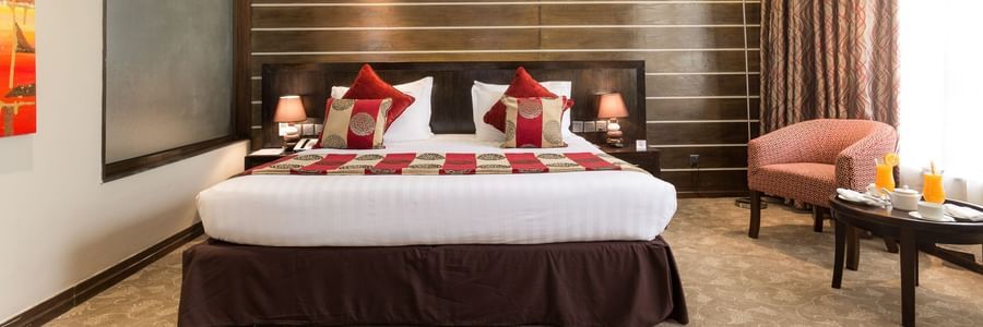 Deluxe Room with king size bed, red cushions, wooden accent wall, armchair with table, carpet flooring, and warm lighting at Boma Inn Eldoret