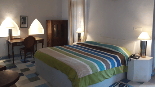 A guest room at Tijara Fort-Palace - 19th Century, Alwar has a bed with striped linens, bedside tables with lamps, a desk with a chair, a wardrobe, an air conditioning unit, and tiled flooring.