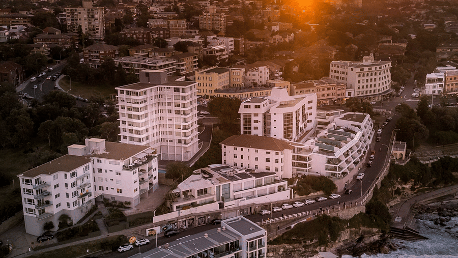 An aerial view of swimming pools at sunrise, with the ocean waves crashing against the pool walls and the city skyline in the background.