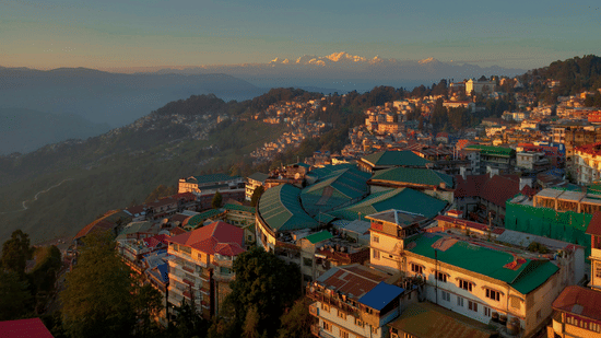 Scenic view of Darjeeling town with colourful hillside buildings, green rooftops, and winding roads, set against misty hills and snow-capped Himalayan peaks under a clear sky at sunrise.