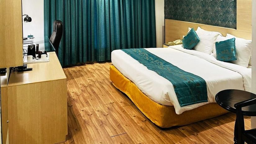 A cosy hotel room at Amer Greens featuring a large bed, hardwood floors, an accent wall, and a large window covered by floor-to-ceiling drapes. Room