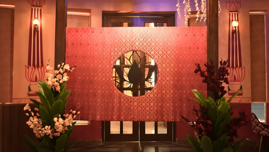 A warmly lit entrance area of Ekam featuring a black and white checkered floor, wooden pillars, a red curtain in the background, flowers, and a chandelier at Kadamb Kuteer, Vrindavan