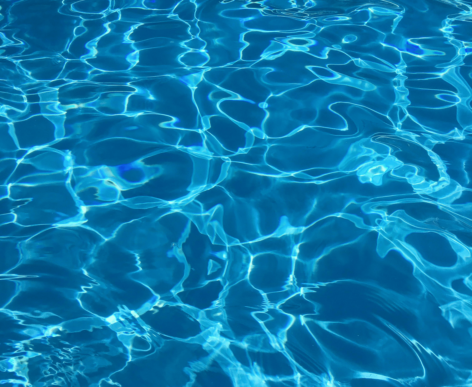 A close-up vertical shot of bright, clear, azure blue water in a swimming pool, with shimmering light reflections on the surface.