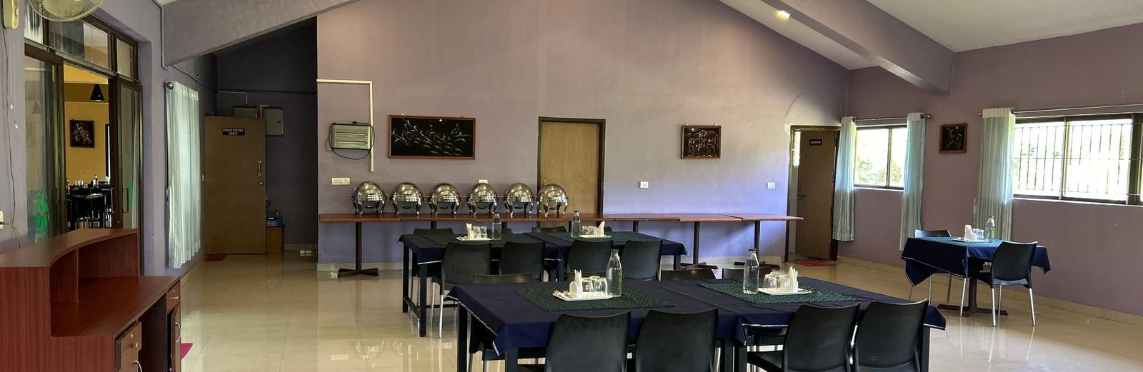 Indoor dining setup with tables, chairs, light purple walls, and ceiling lights at Paradise Wild Hills Resort, Kollur.