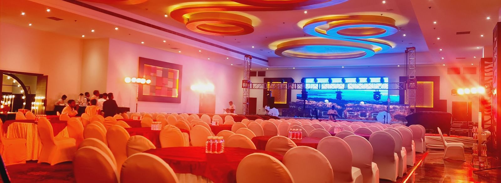 An indoor event space with rows of chairs facing a stage, illuminated by warm lighting and unique circular ceiling fixtures. - Bamboo Saa Resort & Spa, Udaipur