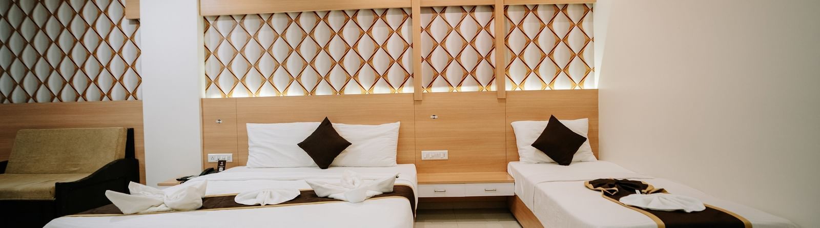 Bright hotel room with a large bed, decorative lattice-patterned wooden headboard, white bedding, and towel art at Daksh The Madhuvan Suites, Dwarka.