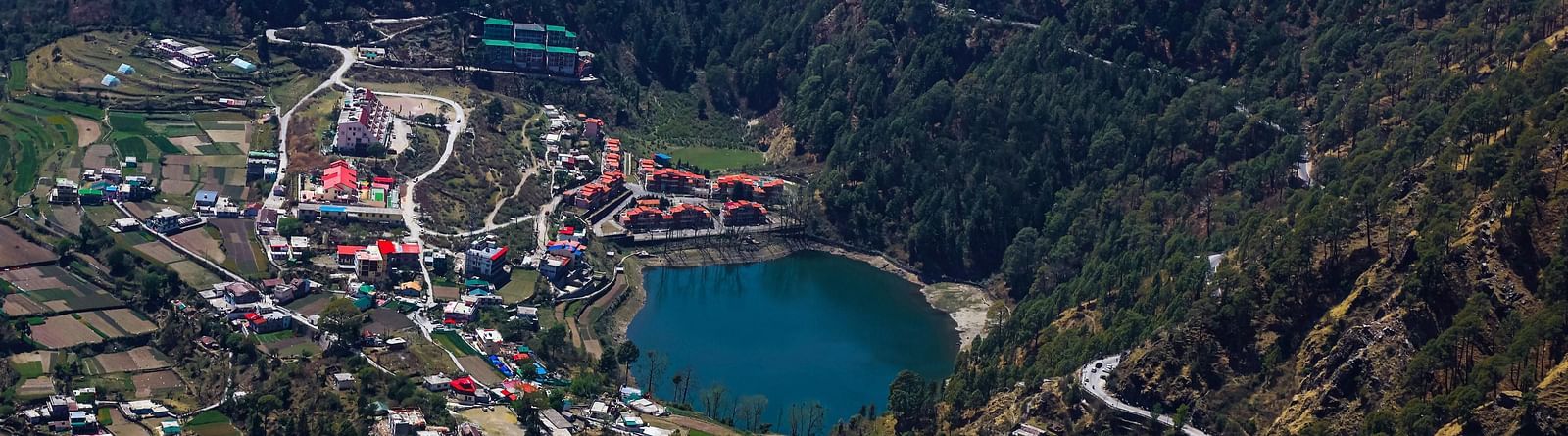 A scenic view of Nainital city  with a lake surrounded by huts and lush green hills in the background.